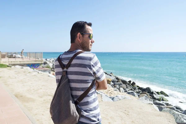 Hombre con bolso de hombro de pie en el paseo marítimo mirando al horizonte en un día soleado — Foto de Stock