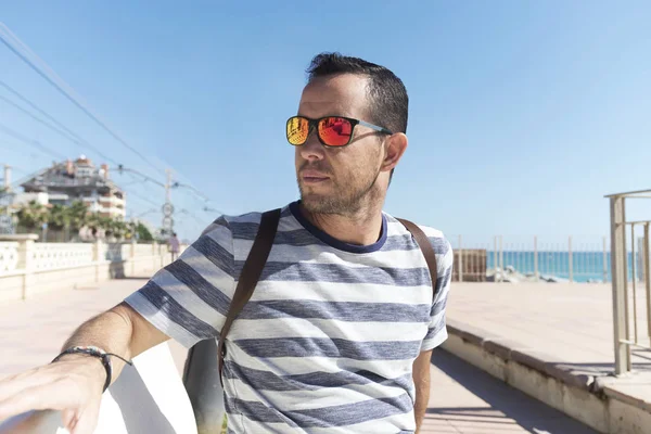 Joven barbudo con gafas de sol sentado en un paseo marítimo mientras mira hacia otro lado — Foto de Stock
