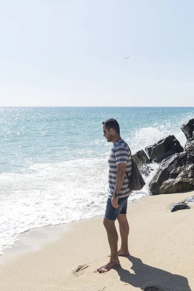 Homem barbudo atraente em pé na costa do mar ao nascer do sol com mochila e horizonte olhando — Fotografia de Stock