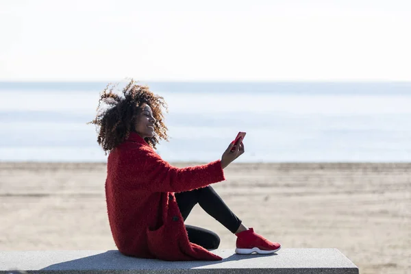 Schöne Afroamerikanerin sitzt an einem sonnigen Tag mit einem Mobiltelefon an Land — Stockfoto