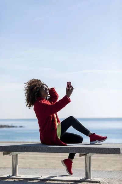 화창 한 날에 핸드폰을 사용하여 해안에 앉아 있는 아름다운아 프로 미국인 여성 — 스톡 사진