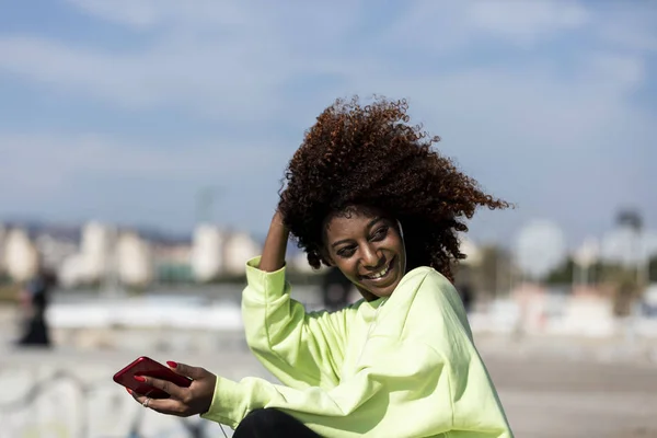 Όμορφη Αφρο-Αμερικανίδα που κάθεται στην ακτή χρησιμοποιώντας ένα κινητό τηλέφωνο σε μια ηλιόλουστη μέρα — Φωτογραφία Αρχείου