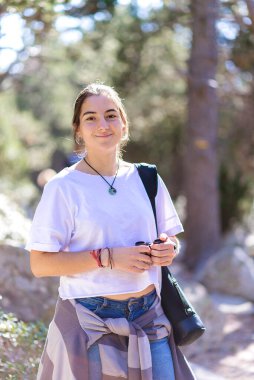 Tişörtlü genç kız arka planda ormanla gülümserken ayakta duruyor.
