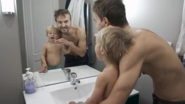 Babası ve küçük oğlu banyoda dişlerini birlikte fırçalıyorlar. Baba, küçük oğlunun dişlerini fırçalamasına yardım ediyor. Aile sabahı rutini. Çocuklara diş bakımı. 