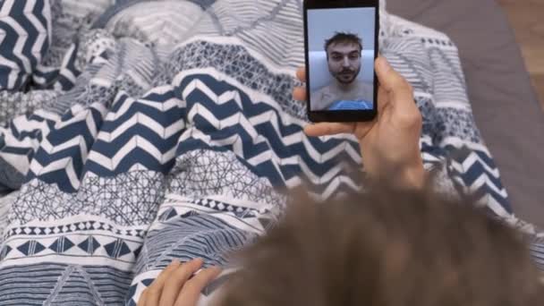 年轻的男性博主使用智能手机躺在床上早起 千年大胡子男子在卧室里制作相声媒体或视频自拍的唱片 — 图库视频影像