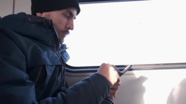Kış parkalı ve örgü şapkalı yakışıklı sakallı adamın yan görünüşü trenle seyahat ederken cep telefonu kullanıyor. Milenyum erkeği toplu taşıma araçlarında akıllı telefonlarda geziniyor ve hapşırıyor.
