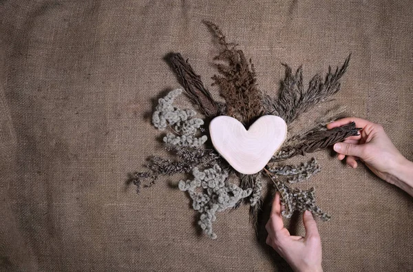 Lasting faded sustainable flowers with wooden heart on natural canvas background. Human hands making composition of dried flowers for Valentine's day on earth tone color burlap. Copy space — Stock Photo, Image