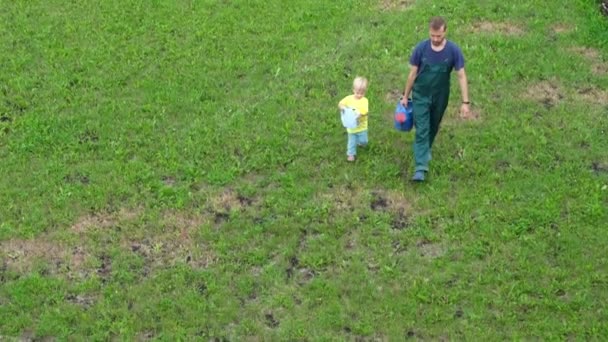 父と息子は緑の芝生の上で水やり缶を手に歩く 庭でお父さんが花に水をやるのを手伝っている子供 上からの眺め — ストック動画