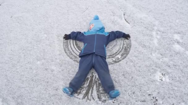 穿着蓝色衣服的小男孩躺在结冰的湖上 成为雪天使 在寒假里玩雪的小男孩真有趣 — 图库视频影像