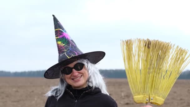 一个身穿女巫服装 戴着圆锥帽 头戴灰色假发 带着现代扫帚的滑稽女人的户外肖像 对着相机笑或笑 万圣节的概念 — 图库视频影像