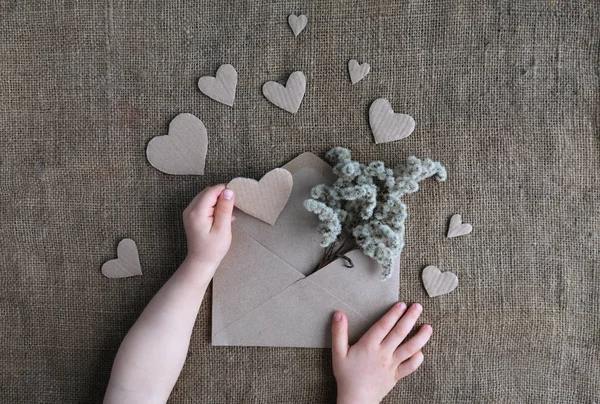 Valentine card in natural earth tone colors. Childs hands holding craft paper envelope with dried flowers and cardboard handmade hearts on canvas burlap background. Valentine day, womans day, love.