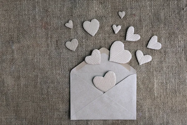 Tarjeta de San Valentín en tonos tierra natural. Sobre de papel artesanal con corazones hechos a mano de cartón sobre fondo de arpillera de lona. San Valentín, día de la mujer, declaración de amor, invitación a la boda . — Foto de Stock