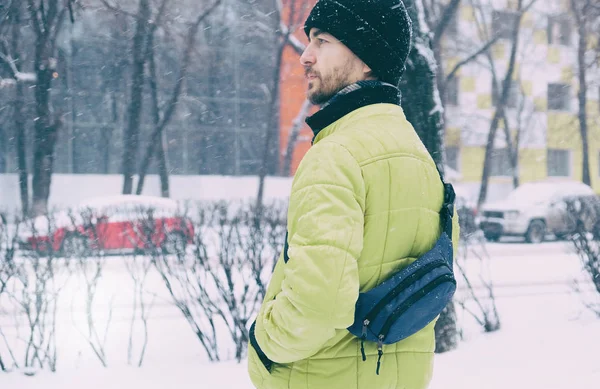 Extremo inverno tempestade de neve nevasca do tempo na cidade. Casual homem milenar em amarelo jaqueta inchada e chapéu de malha com saco crossbody stand na rua nevada . — Fotografia de Stock