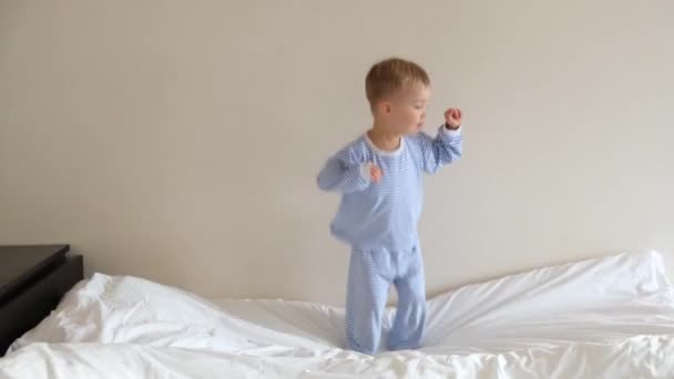 ベッドの上でジャンプ青い縞のパジャマでかわいい幼児の男の子の子供 笑顔と歌 幸せな子供時代のコンセプト — ストック動画
