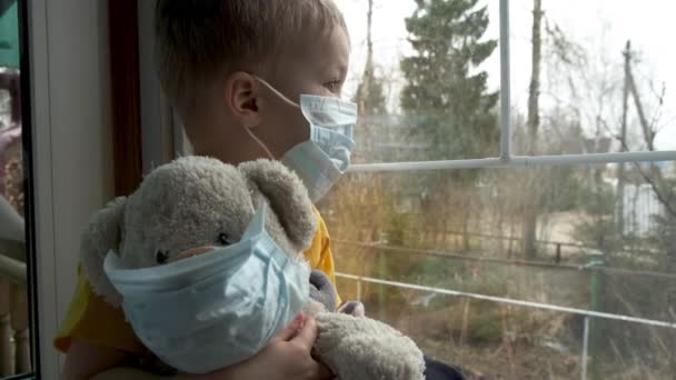 家庭内隔離の病気の子供 男の子とテディは両方とも防護医療マスクで窓辺に座って窓の外を見る ウイルスの保護 コロナウイルスのパンデミック 予防流行 — ストック動画