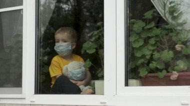 Ev karantinasında sevimli bir çocuk. Koruyucu tıbbi maskeli oyuncak ayılı çocuk pencere pervazında oturur ve pencereden dışarı bakar. Alerji, covid-19 koronavirüs salgını, önleme salgını.
