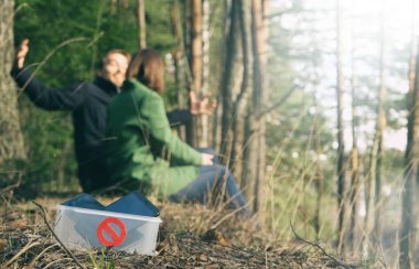 Dijital detoks konsepti. Çiftler doğada akıllı telefonlar olmadan zaman geçirirler. Kadın ve erkek parkta oturuyor, konuşuyor ve gülüyor, telefonlar kutuda ve ön planda yasak işareti var.. 