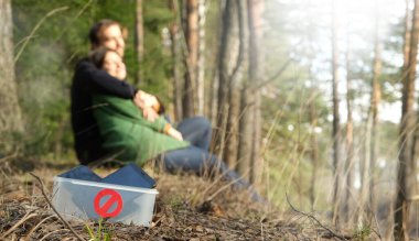 Dijital detoks konsepti. Çiftler doğada akıllı telefonlar olmadan zaman geçirirler. Erkek ve kadın parkta ya da ormanda oturuyor, sarılıyorlar, telefonlar kutuda ve ön planda yasak işareti var. Pankart.
