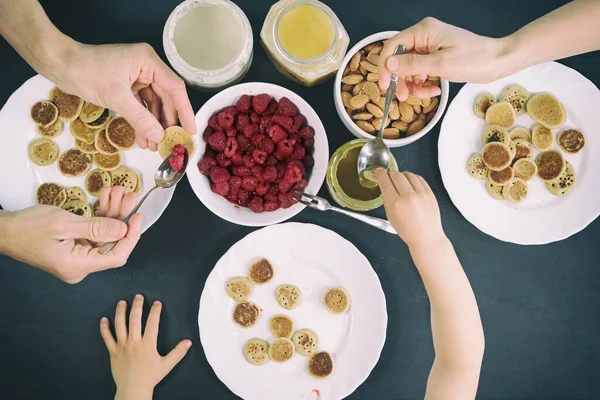 Top view family eating homemade breakfast. Tiny mini pancake cereal, sweet spreads, nuts, honey and fresh raspberries. Parents and child eat trendy quarantine food. Black table with meal flat lay.