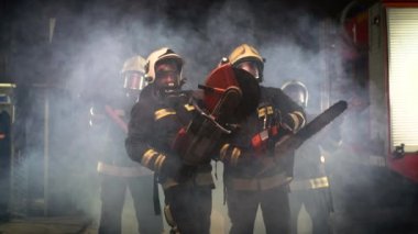 Tam teçhizatlı itfaiyeciler, oksijen maskeleri ve acil durum kurtarma araçları, dairesel hidrolik ve gaz testeresi, balta ve balta çekici. Arka planda duman ve itfaiye araçları. 