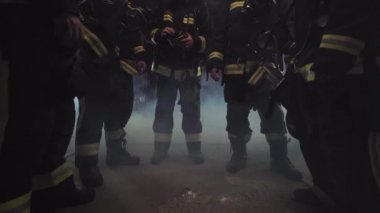 Tam teçhizatlı beş itfaiyeci grubu. İş bittikten sonra gevşemiş sohbet. Arka planda duman ve itfaiye araçları var. İsim etiketi çevirisi itfaiyeci ve modelin soyadı.