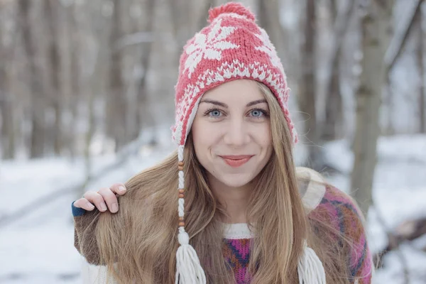 冬の肖像を着た若い美しい金髪女性のニット帽子です。雪が降る冬美容ファッションのコンセプトです。背景の雪に覆われた森. — ストック写真
