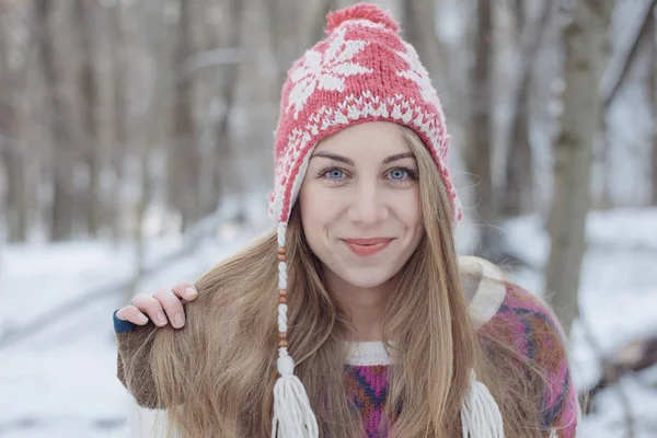 冬の肖像を着た若い美しい金髪女性のニット帽子です。雪が降る冬美容ファッションのコンセプトです。背景の雪に覆われた森. — ストック写真