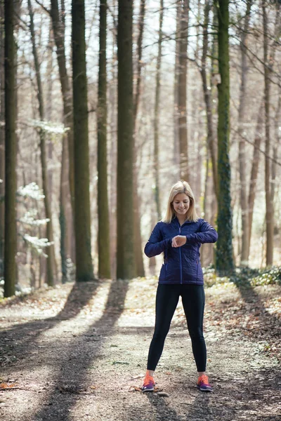 Trail running runner looking at heart rate monitor watch running in forest wearing warm jacket sportswear. Female jogger running training in woods. — Stock Photo, Image
