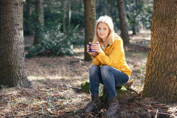 Carino donna viaggiatore bionda a riposo nella foresta e bere caffè caldo o tee da una tazza vintage — Foto Stock