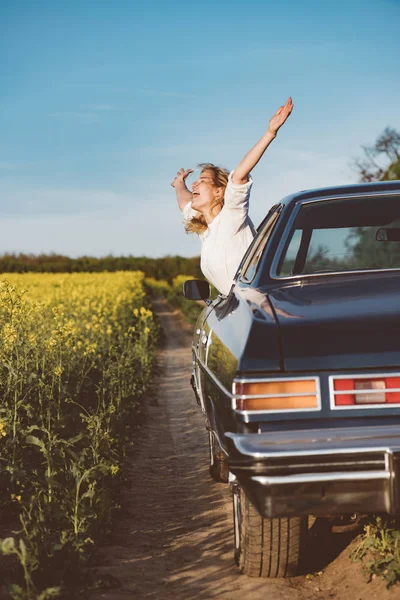 Путешествует с удовольствием. Богатая молодая женщина ликует от волнения, высунувшись из окна машины с протянутыми руками, празднуя начало летних каникул. — стоковое фото