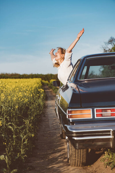 Traveling with fun. Exuberant young woman cheering with excitement as she leans out of the car window with outstretched arms celebrating the start of her summer vacation