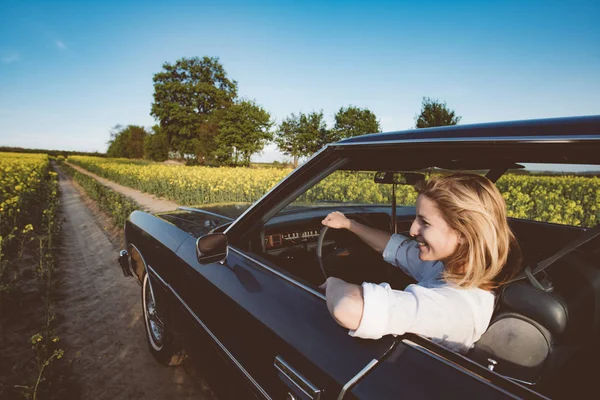 Giovane pilota donna alla guida di una grande muscle car americana V8 su strada aperta. Viaggio su strada, viaggio con auto d'epoca . — Foto Stock