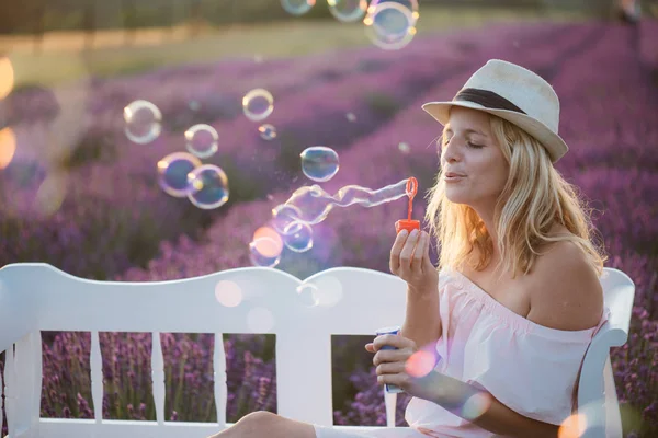 Krásná žena, foukání mýdlové bubliny na lavičce. Na pozadí nádherné levandulové pole. — Stock fotografie