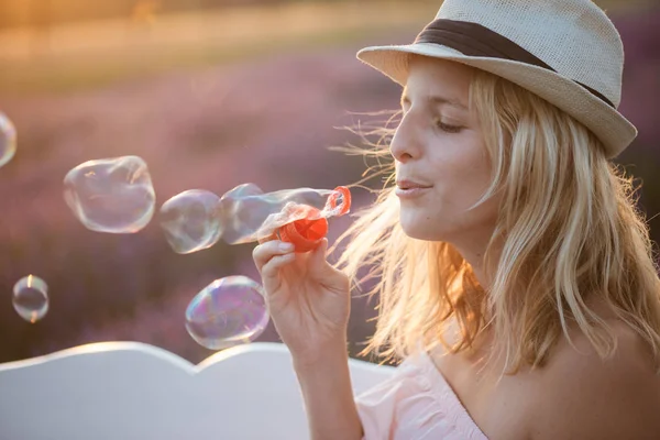 Bella donna che soffia bolle di sapone su una panchina. Magnifico campo di lavanda sullo sfondo . — Foto Stock