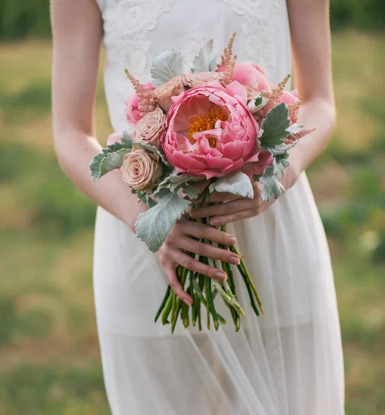 Panna Młoda w sukni, stojąc w zielonym ogrodzie i trzymając Bukiet ślubny, kwiaty i zieleń. Kobieta trzyma kolorowy bukiet z nią ręce na dzień ślubu — Zdjęcie stockowe