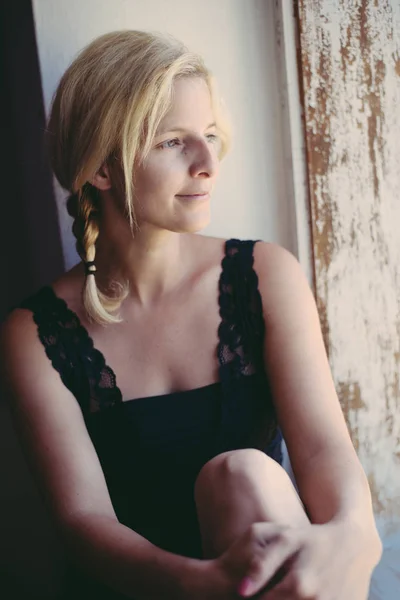 Woman portrait at home. Long blonde hair and pigtail. Female looking out of window. — Stock Photo, Image