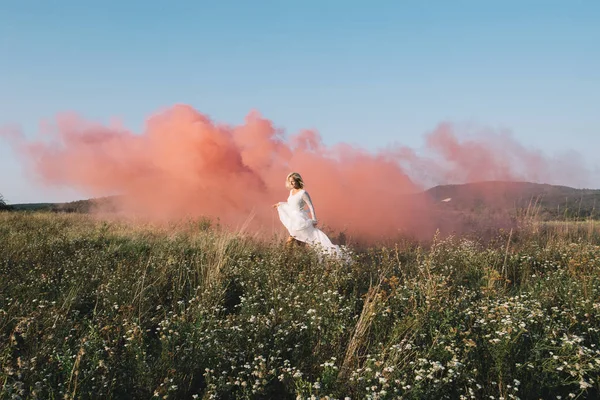 Novia en vestido de novia blanco caminando con humo rojo — Foto de Stock