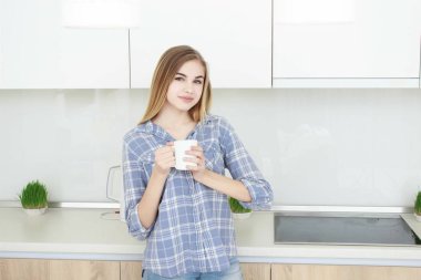 Genç güzel kız mavi kareli gömlek ve kot Mutfakta çay veya kahve ile beyaz bir bardak içme. Sabah. ev iç.
