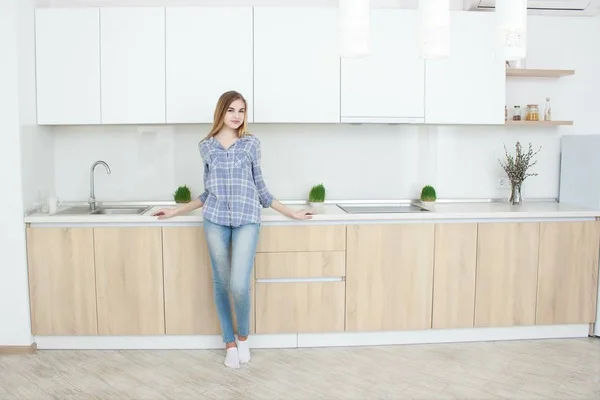 V kuchyni stojí krásná mladá dívka v modré kostkované košili a modré džíny. Domácí interiér — Stock fotografie