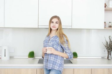 Genç güzel kız mavi kareli gömlek ve kot Mutfakta çay veya kahve ile beyaz bir bardak içme. Sabah. ev iç.
