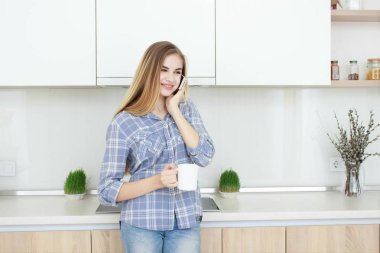 Genç güzel kız mavi kareli gömlek ve kot Mutfakta çay veya kahve ile beyaz bir bardak içme. Sabah. ev iç.