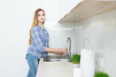 Genç güzel kız mavi kareli gömlek ve kot mutfakta bardak su dökme. Sabah. ev iç.