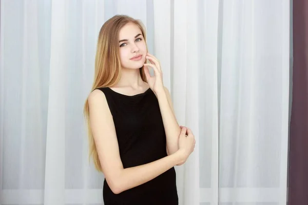 Young girl with black dress against curtains. Looking at the camera. — Stock Photo, Image