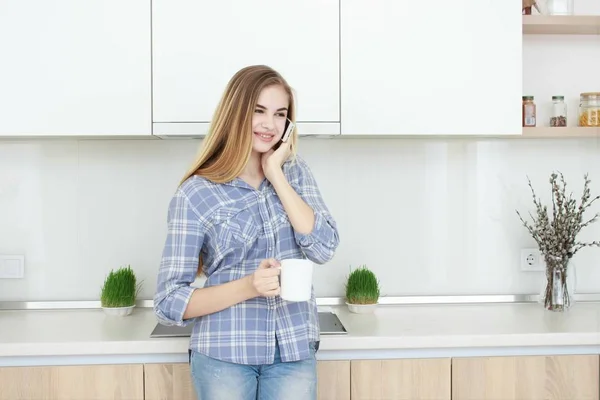 Молодая красивая девушка в голубой клетчатой рубашке и джинсах на кухне пьет чай или кофе с белой чашкой. Доброе утро. домашний интерьер . — стоковое фото