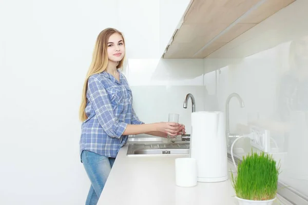 Молодая красивая девушка в голубой клетчатой рубашке и джинсах на кухне наливает стакан воды. Доброе утро. домашний интерьер . — стоковое фото