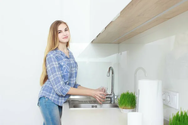Молодая красивая девушка в голубой клетчатой рубашке и джинсах на кухне наливает стакан воды. Доброе утро. домашний интерьер . — стоковое фото