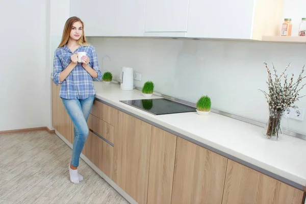 Young beautiful girl in a blue checkered shirt and jeans in the kitchen drinking tea or coffee with a white cup. Morning. home interior. — Stock Photo, Image