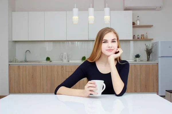 Frau lächelnd in Küche mit Kaffeetasse sitzend. schönes Mädchen mit langen Haaren. — Stockfoto