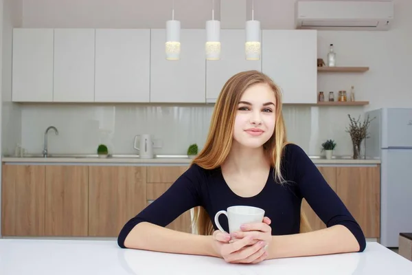 Frau lächelnd in Küche mit Kaffeetasse sitzend. schönes Mädchen mit langen Haaren. — Stockfoto
