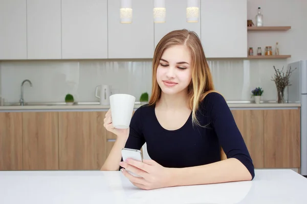 Belle fille en vêtements de maison utilise un smartphone, boire du thé et sourire tout en étant assis dans la cuisine — Photo